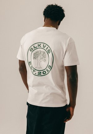 Mann mit kurzen lockigen Haaren trägt weißes T-Shirt mit grünem kreisförmigem Logo, das einen Baum und den Text "BLKVIS EST. 2013" auf dem Rücken zeigt, steht vor einfachem Hintergrund.
