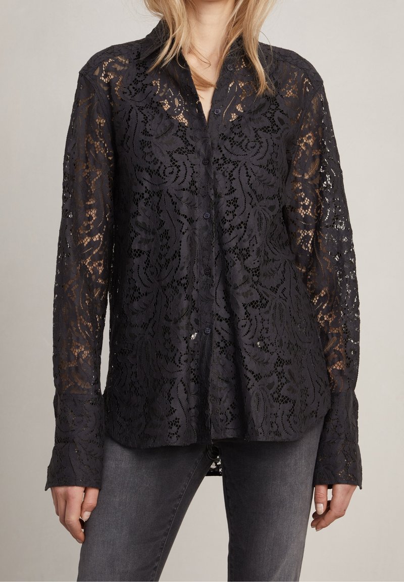 Camicia nera in pizzo con motivo floreale, maniche lunghe e design con bottoni. Il tessuto è semi-trasparente con una finitura testurizzata.