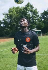 Svart Nike fotballdrakt med prikkete mønster, oransje løveemblem og hvit designelement. Modellen ser opp på en fotball på en gresslette.