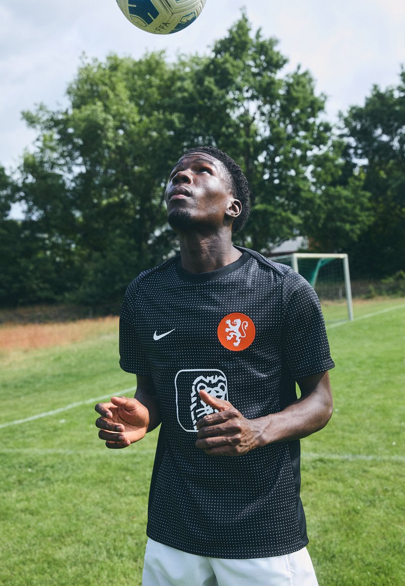 Svart Nike fotballdrakt med prikkete mønster, oransje løveemblem og hvit designelement. Modellen ser opp på en fotball på en gresslette.