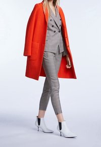 Manteau en laine orange vif par-dessus un costume à carreaux gris, pantalon slim et bottines blanches avec des accents noirs.