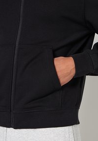 Czarna bluza z kapturem zapinana na zamek, o gładkiej fakturze, z przednią kieszenią i ściągaczami na mankietach. Na zdjęciu widoczna ręka spoczywająca w kieszeni.