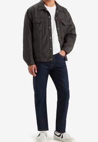 Chaqueta acolchada de color gris oscuro con cierre de botones a presión, dos bolsillos en el pecho y un collar, combinada con jeans oscuros y zapatillas blancas.