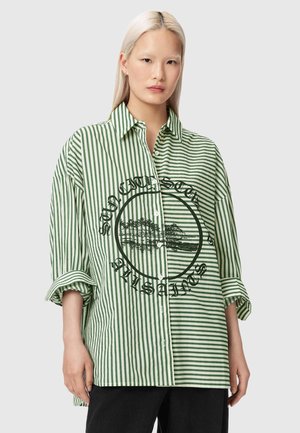 Jeune femme aux longs cheveux blonds portant une chemise à boutons oversize à rayures verticales vertes et blanches avec un motif noir et des manches retroussées.