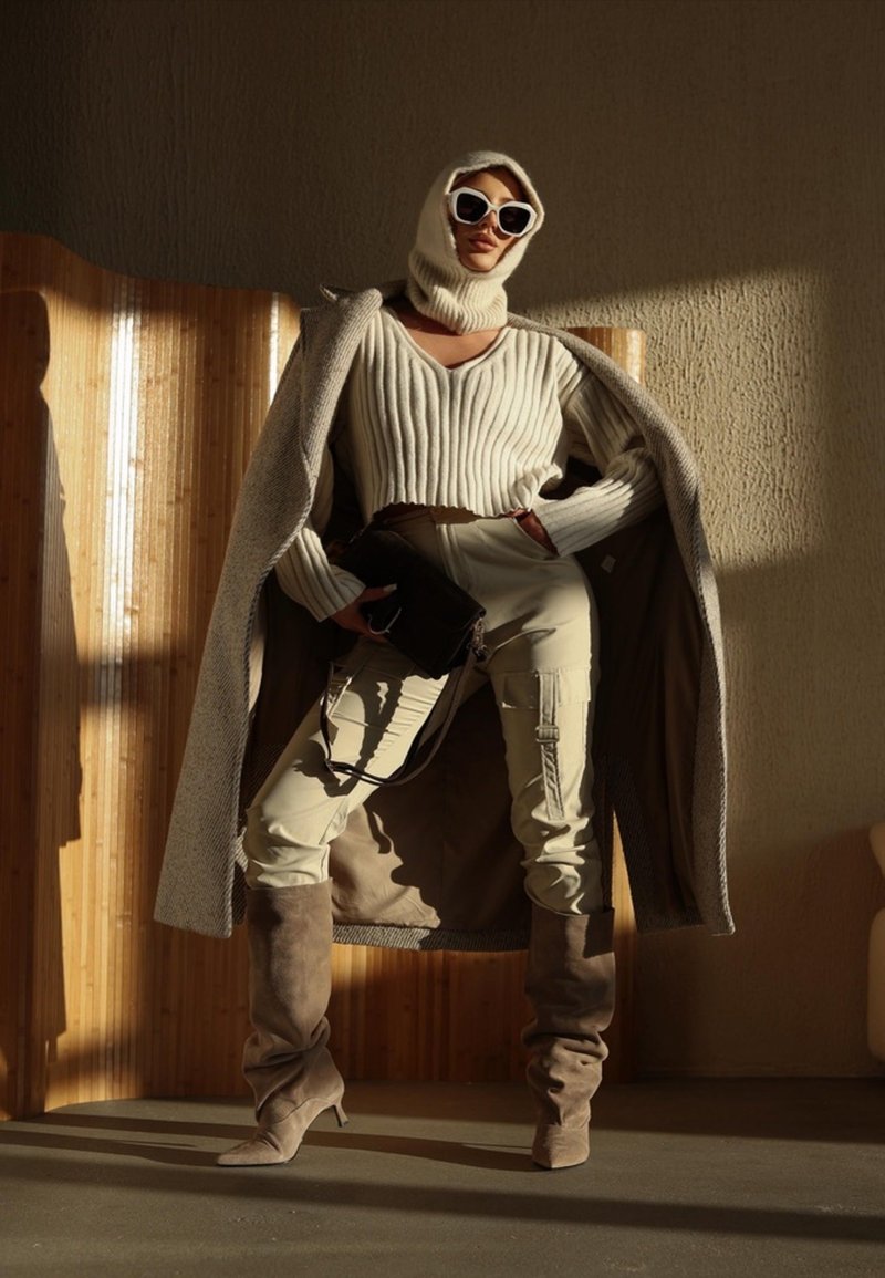 Vrouw met beige geribbelde trui, cargobroek, losse laarzen, oversized jas over de schouders gedrapeerd, gebreide capuchon, witte zonnebril, zwarte tas in de hand.
