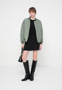 Giacca bomber verde oliva, top aderente nero, minigonna nera, stivali neri di pelle alto fino al ginocchio, piccola borsa nera con manici.