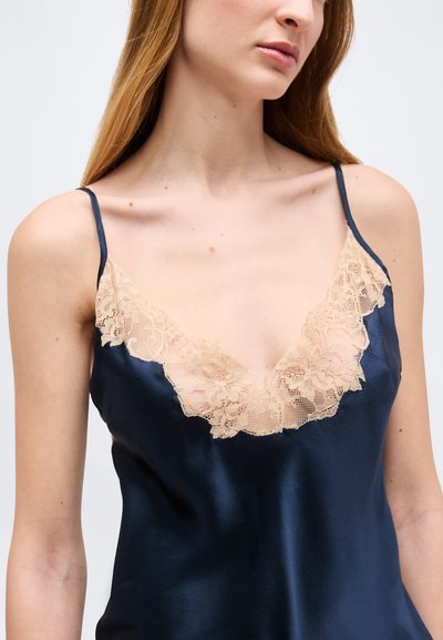 Vrouw draagt een marineblauwe satijnen camisole met beige kanten rand en dunne schouderbandjes, weergegeven van nek tot taille tegen een witte achtergrond.