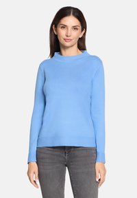 Kobieta o prostych brązowych włosach ubrana w jasnoniebieski sweter z długim rękawem i szare dżinsy, stojąca na tle jednolitej białej ściany.