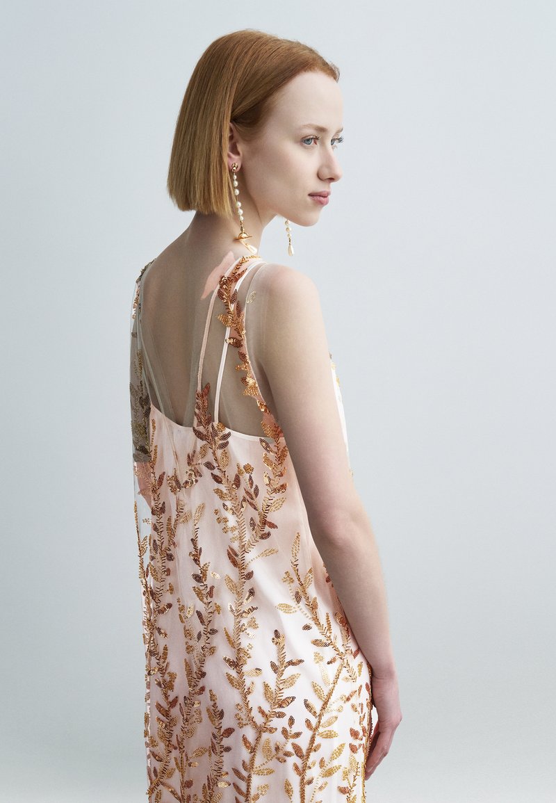 Ένα αμάνικο φόρεμα σε απαλό ροζ ύφασμα, με χρυσή κεντητική με φύλλα και διάφανα πλαϊνά, συνοδευόμενο από μακριά κρεμαστά σκουλαρίκια που προσθέτουν λεπτομέρεια.