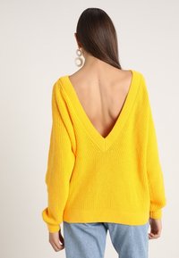 Żółty dziergany sweter z głębokim dekoltem w kształcie litery V z tyłu, długimi rękawami i ściągaczami przy mankietach. Neutralne tło, aby skupić uwagę na szczegółach ubrania.