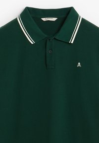 Polo de color verde oscuro fabricado con tejido suave. Presenta un cuello clásico con rayas blancas y un pequeño logo de calavera en el lado izquierdo del pecho.