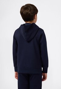 Ragazzo in piedi con le spalle alla macchina fotografica, indossa una felpa blu navy semplice e pantaloni blu navy abbinati su uno sfondo bianco.