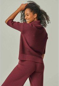 Burgunderfarbener, gerippter Strickpullover mit hohem Kragen und kurzen Ärmeln; kombiniert mit passenden, weit geschnittenen Hosen, die einen lockeren Schnitt und eine weiche Textur aufweisen.