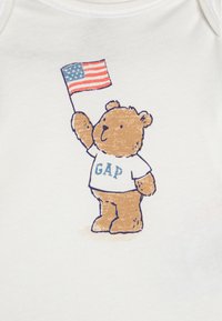 Βρεφικό φορμάκι σε λευκό χρώμα με γραφικό ενός καφέ αρκούδου που κρατά μια μικρή αμερικανική σημαία, φορώντας ένα μπλουζάκι που λέει "GAP" σε μπλε κείμενο.