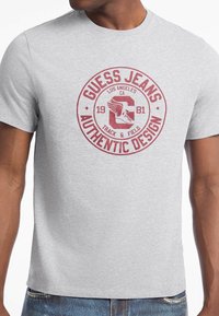 Grå bomulds T-shirt med et rundt rødt grafisk logo "GUESS JEANS AUTHENTIC DESIGN" og "TRACK & FIELD," med et vingeemblem.