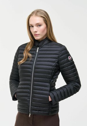 Donna che indossa una giacca nera imbottita con zip e toppa logo sulla manica, mani nelle tasche, su sfondo chiaro semplice.