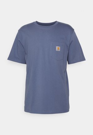 Personne portant un t-shirt Carhartt bleu et un pantalon noir assise sur une chaise en grille métallique bleue, montrant des chaussettes blanches et des baskets Adidas bleues.