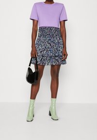 Fialové zkrácené tričko, černá sukně s květinovým vzorem a volány, zelené lakované kožené kotníkové boty, držící černou kabelku.