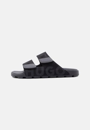Black slide sandal with two adjustable gray straps, textured sole, and subtle embossed "HUGO" logo on the side.
