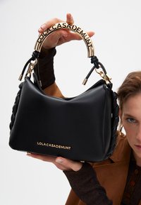 Una persona tiene in mano una borsa nera con un design intrecciato e un manico con logo dorato, mostrando la borsa su uno sfondo neutro.