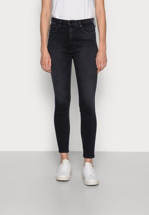 Persona que lleva vaqueros pitillo negros, una camiseta blanca y zapatillas blancas, de pie sobre un fondo blanco liso.