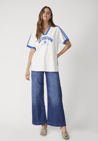 T-shirt bianca oversized a scollo a V con dettagli blu e testo, abbinata a jeans larghi in denim scuro. Le calzature sono chiare e chunky.