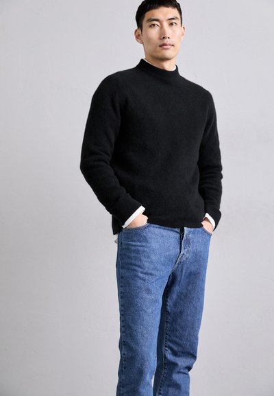 Μαύρο πλεκτό πουλόβερ με ψηλό λαιμό, συνδυασμένο με ανοιχτό μπλε τζιν. Τα μανίκια του πουλόβερ διαθέτουν λευκές λεπτομέρειες.
