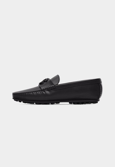 Mocassin en cuir noir avec semelle en caoutchouc à profil bas et clous, orné d'un pompon décoratif sur le dessus, présenté en vue latérale sur fond uni.