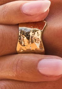 Goldfarbener Ring mit einem wellenförmigen, unregelmäßigen Design und einer polierten Oberfläche. Besitzt sichtbare Texturen und Reflexionen und wird gegen die Haut gehalten.