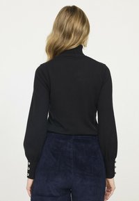 Pull à col roulé noir à manches longues. Présente un tissu texturé et des boutons décoratifs au niveau des poignets. Porté avec un pantalon en velours côtelé bleu marine.