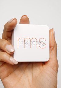 Vit kompakt med rundade kanter, med texten "rms beauty" i grått och klargult. Hållen i en hand med fina, ljusa naglar.