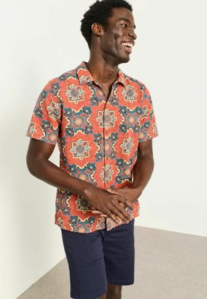 Lächelnder Mann, der ein kurzärmeliges, gemustertes orangefarbenes Hemd mit Knöpfen und marineblaue Shorts trägt, steht mit gefalteten Händen.