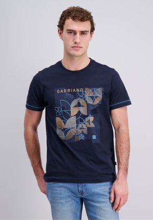 Jonge man draagt een donkerblauw Gabbiano t-shirt met een blauw en bruin geometrisch bladvormig patroon, gecombineerd met lichtblauwe jeans.