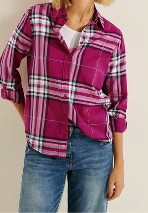 Femme portant une chemise à carreaux magenta aux manches retroussées sur un top blanc avec un jean bleu, debout devant un fond neutre.