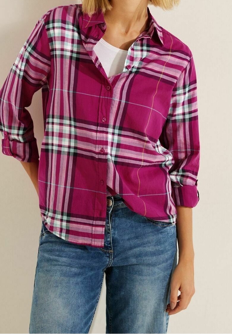 Femme portant une chemise à carreaux magenta aux manches retroussées sur un top blanc avec un jean bleu, debout devant un fond neutre.