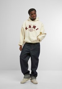 Felpa color crema con il testo "KANI" in bordeaux, abbinata a jeans larghi blu scuro e sneakers beige. Tessuti morbidi e vestibilità casual.
