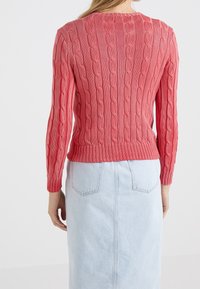 Pull rose en maille torsadée avec manches longues et encolure arrondie, présentant des motifs texturés ainsi que des poignets et un ourlet côtelés. Associé à une jupe en jean.