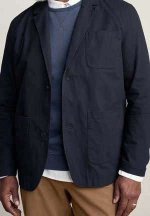 Mand iført marineblazer over blå sweater og hvid skjorte med brune bukser, stående med hænderne synlige langs siden.