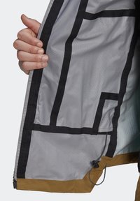 Main tenant une veste ouverte montrant la doublure intérieure avec des coutures noires, une bordure marron et un cordon de serrage ajustable à l'ourlet.