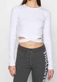 Vit ribbad långärmad croptop med ett korsat framdesign, tillsammans med grå träningsbyxor med vitt logodetalj.