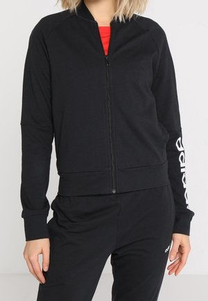Femme portant une veste noire zippée avec un texte blanc sur la manche et un pantalon noir assorti sur un fond uni.