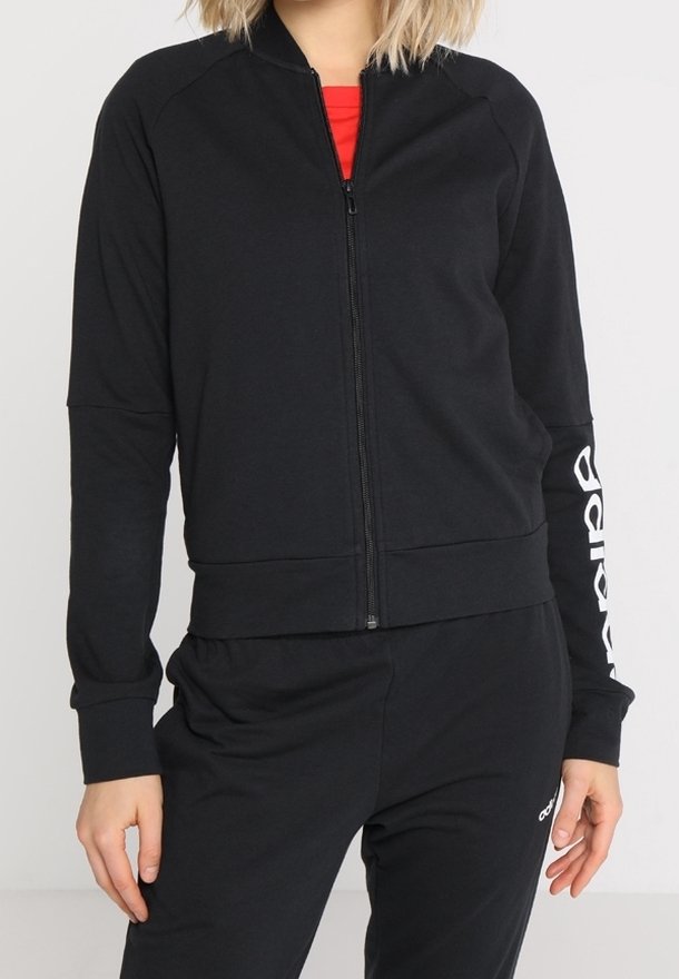 Femme portant une veste noire zippée avec un texte blanc sur la manche et un pantalon noir assorti sur un fond uni.