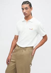 Witte polo met kraag, voorzien van een klein rood en geel logo. Gecombineerd met beige, geplooide broek voor een casual look.