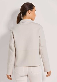 Frau mit dunklem Haar, die einen Pferdeschwanz trägt, bekleidet mit einer hellbeigen Langarmjacke und passenden Hosen, die von hinten vor einem schlichten Hintergrund steht.