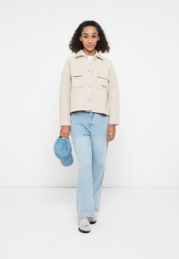 Beige cropped jacket with two front pockets, button closure, paired with light blue wide-leg jeans and a blue cap, set against a white background.
