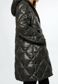 Donna che indossa un cappotto imbottito nero a quadretti con cappuccio, mani nelle tasche laterali, mostrata di profilo contro uno sfondo neutro.