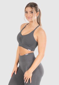 Jonge vrouw met lang haar in een paardenstaart, gekleed in een grijze sport-bh en hoge taille leggings, die glimlacht en poseert tegen een effen achtergrond.