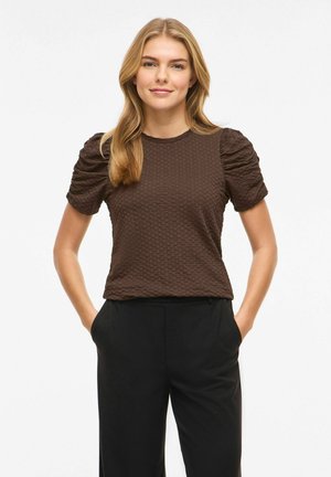 Jeune femme aux longs cheveux blonds portant un haut marron texturé à manches courtes et un pantalon noir, debout les mains dans les poches sur fond blanc.