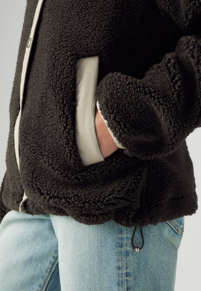 Veste en polaire noire avec accents crème, présentant une texture à poils longs, poches latérales, cordon élastique à la base et fermeture à boutons.