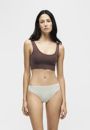 Femme aux longs cheveux foncés portant un crop top marron et des sous-vêtements beige clair, debout devant un fond blanc uni.